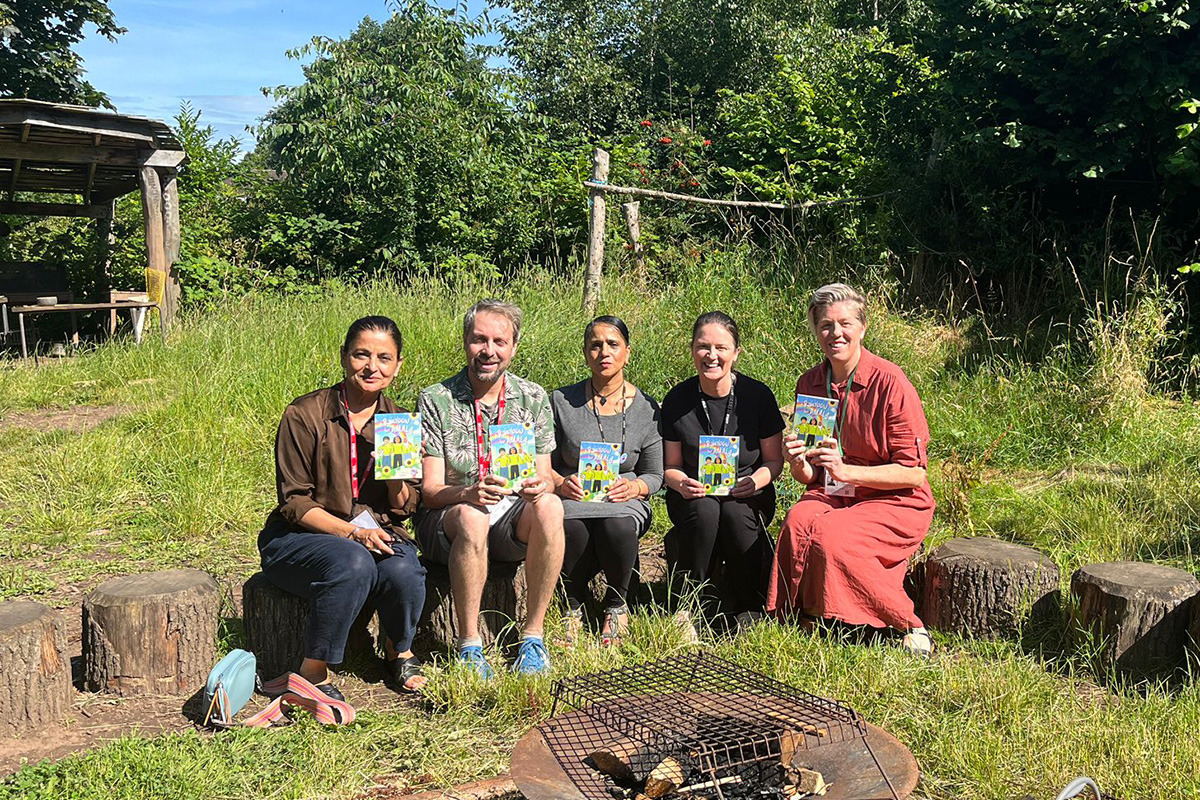 A group of five adults sitting on logs in a community garden. They are holding copies of the book A Rainbow for Amala.