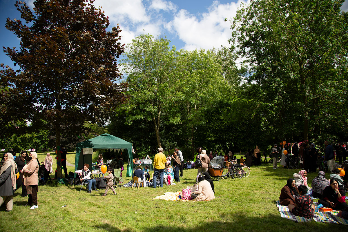 A gathering of people in a grassy clearing in a woodland. There is a small gazebo.