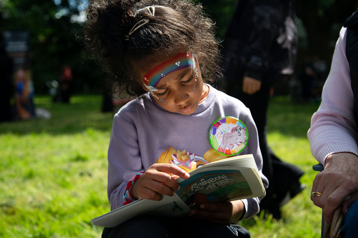A young person with rainbow face paint is reading a book on sunlit grass.
