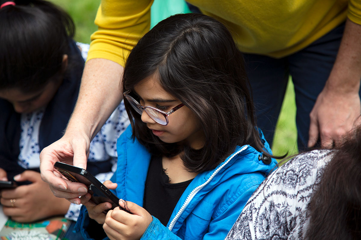 A young person with shoulder length hair and glasses is looking intently at a mobile phone held by an adult.