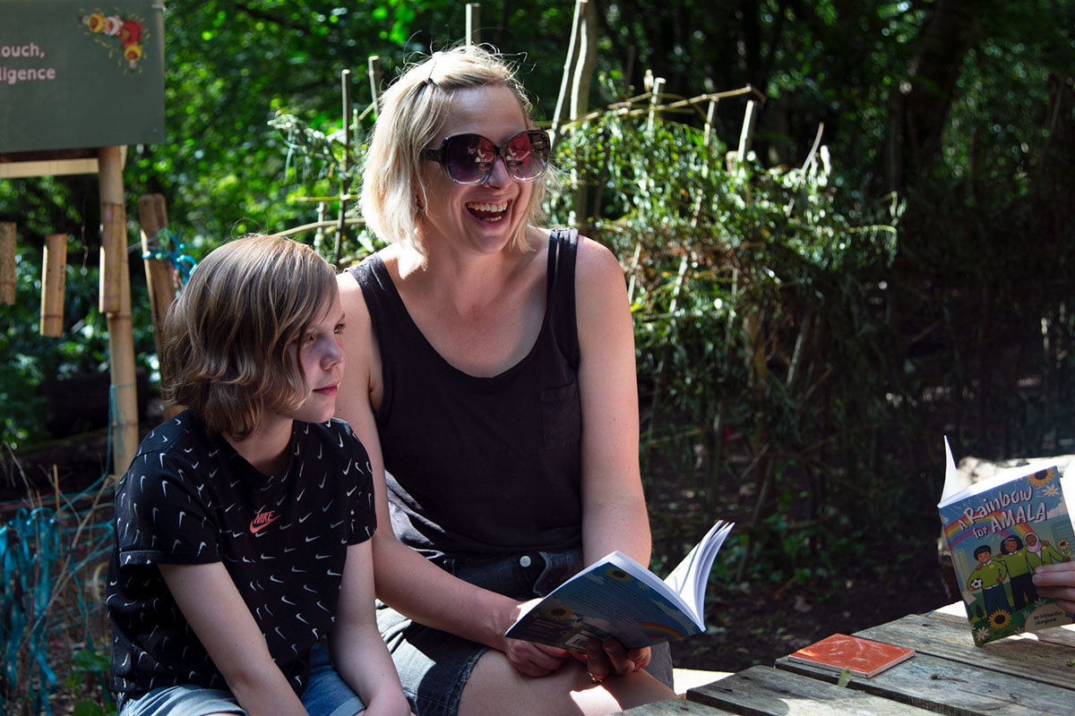 An adult in sunglasses laughts as she reads a book to a young person.
